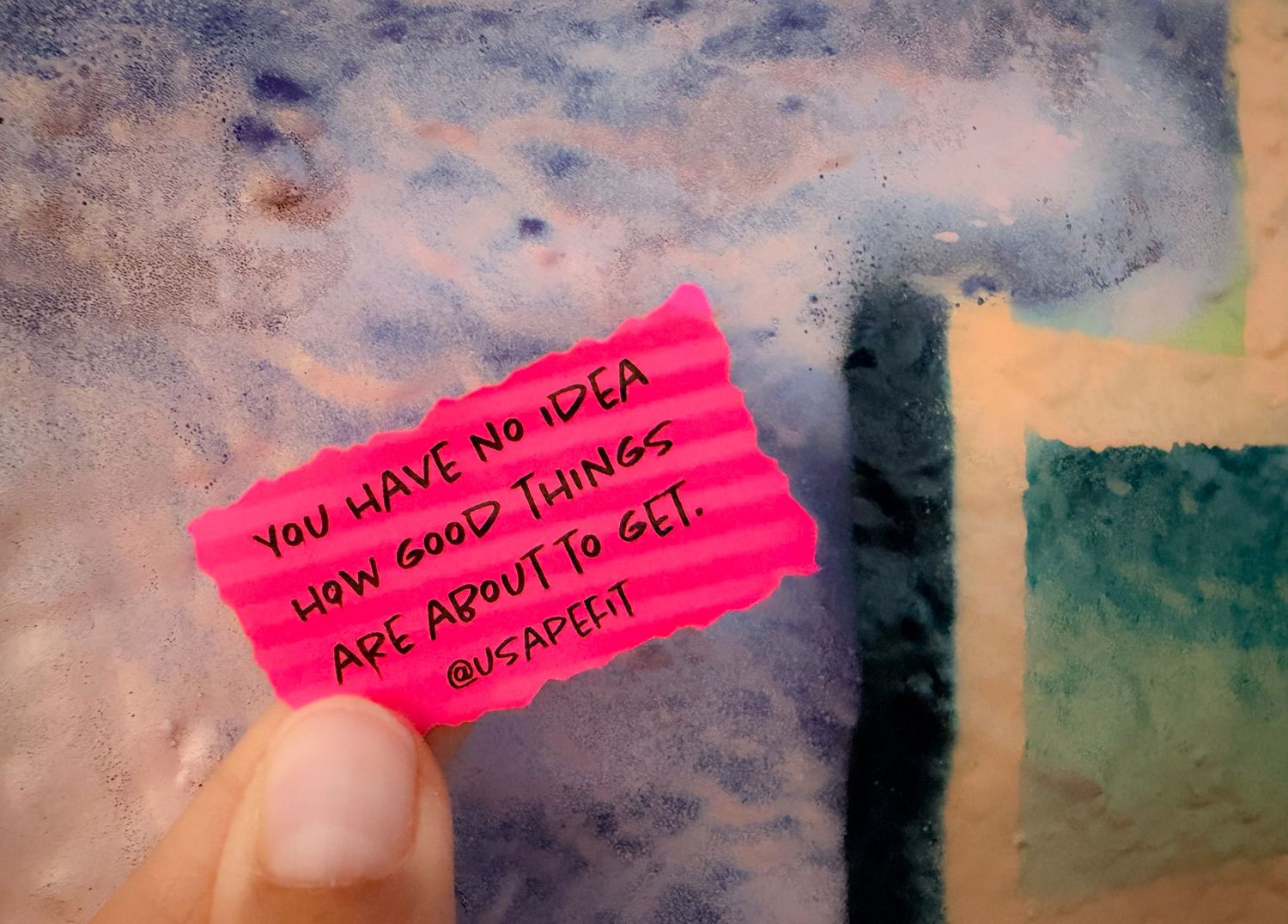 Hand holding a bright pink note card with motivational quote about good things coming, against a blue abstract background.