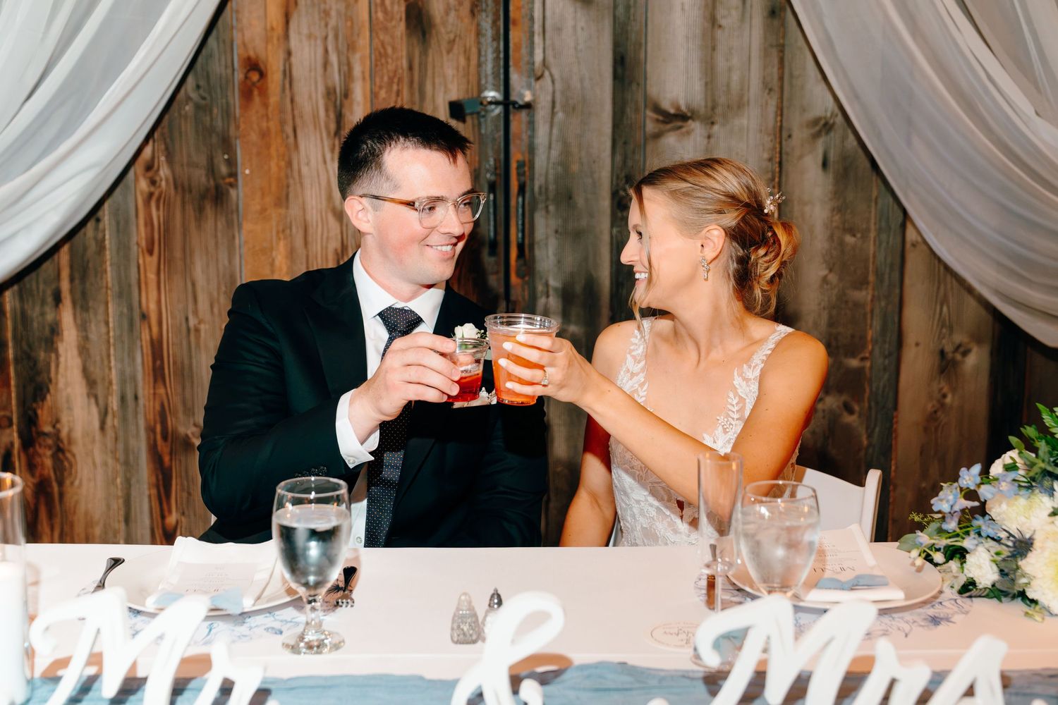 Bride and groom toasting drinks at wedding reception table with Mr. and Mrs. signs in rustic barn venue.
