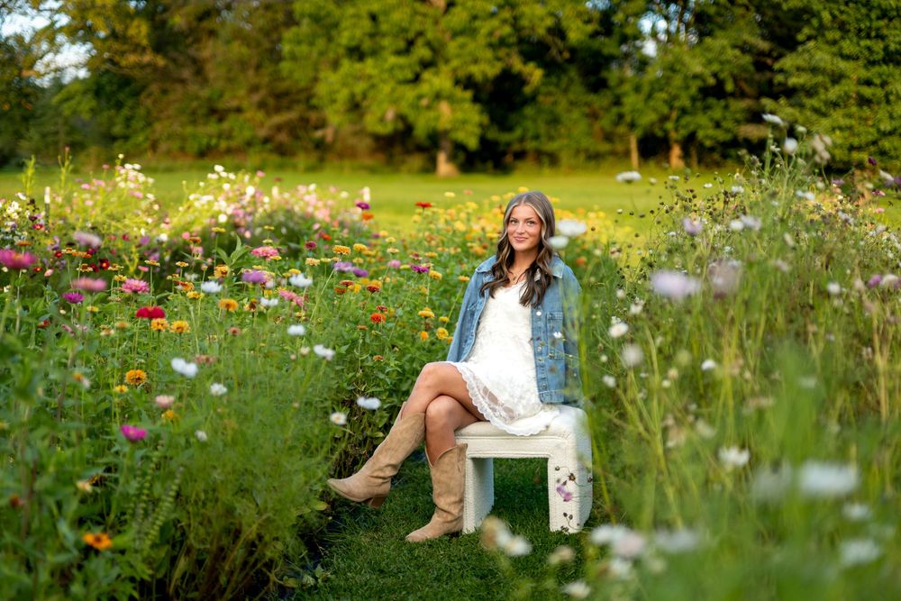 Young woman in white dress and denim jacket sits on white chair surrounded by colorful wildflowers in lush green garden.