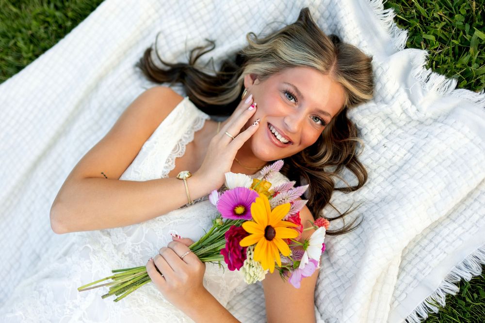 Young woman lying on white blanket outdoors, smiling and holding colorful flower bouquet with pink and yellow blooms.