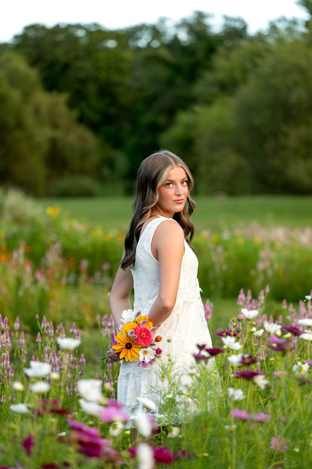 Young woman in white dress holding colorful bouquet stands in vibrant wildflower meadow with lush green trees behind her.