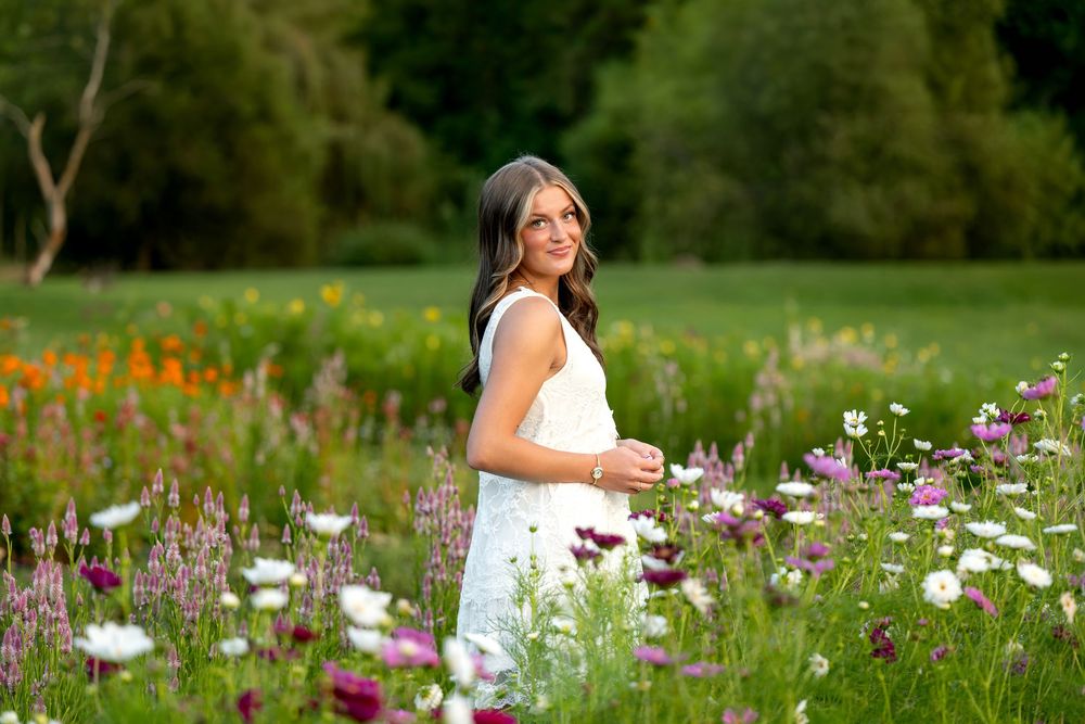 A smiling woman in a white dress stands among colorful wildflowers in a lush green meadow.