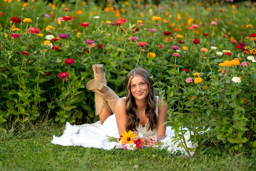 Young woman in white dress lying in a vibrant flower field with colorful zinnias blooming behind her.