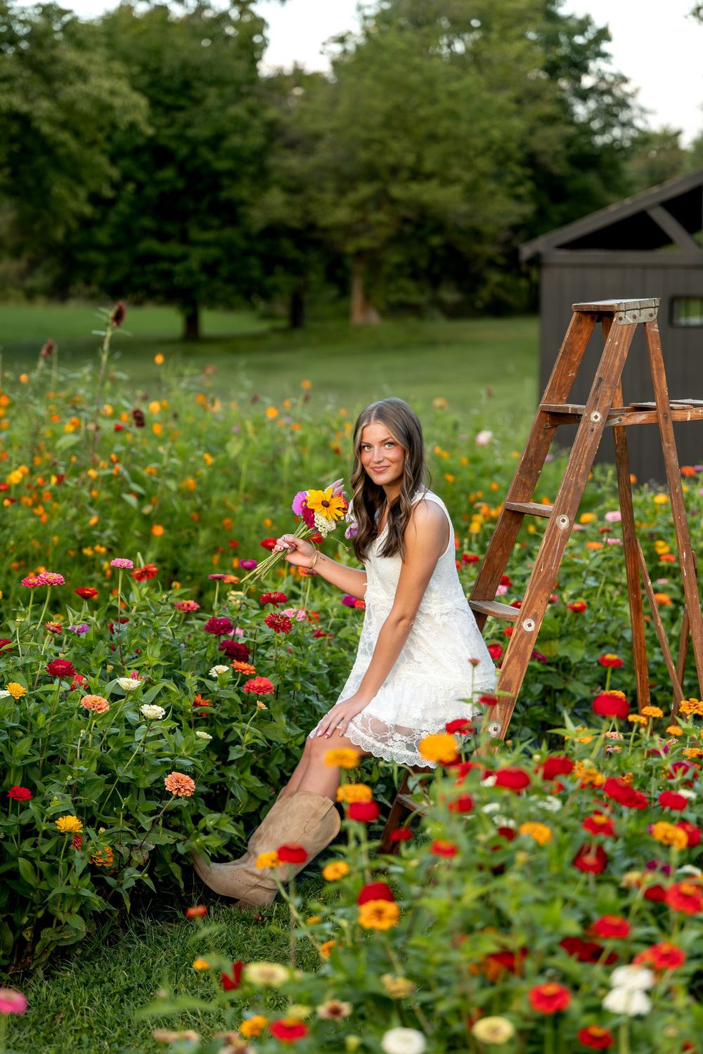 Young woman in white lace dress and cowboy boots holding flowers beside a wooden ladder in a colorful zinnia garden.