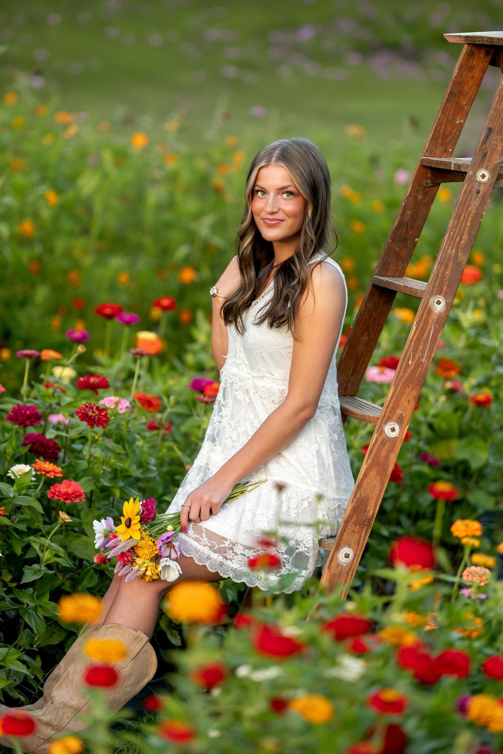Smiling brunette woman in white dress leaning against rustic wooden ladder surrounded by vibrant multicolored zinnias.