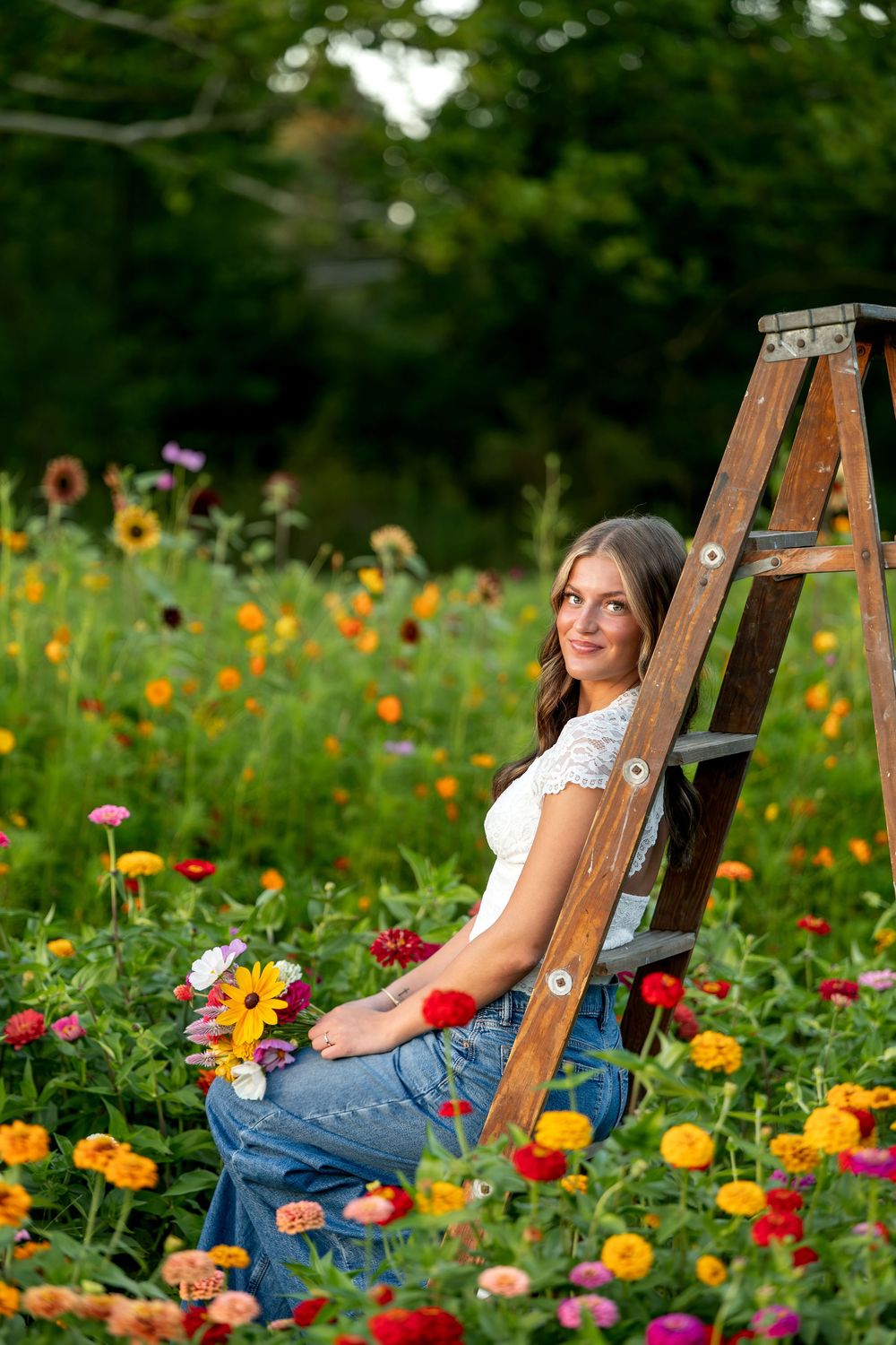 Young woman in white lace top and jeans sitting among vibrant zinnias leaning against a wooden ladder in garden.