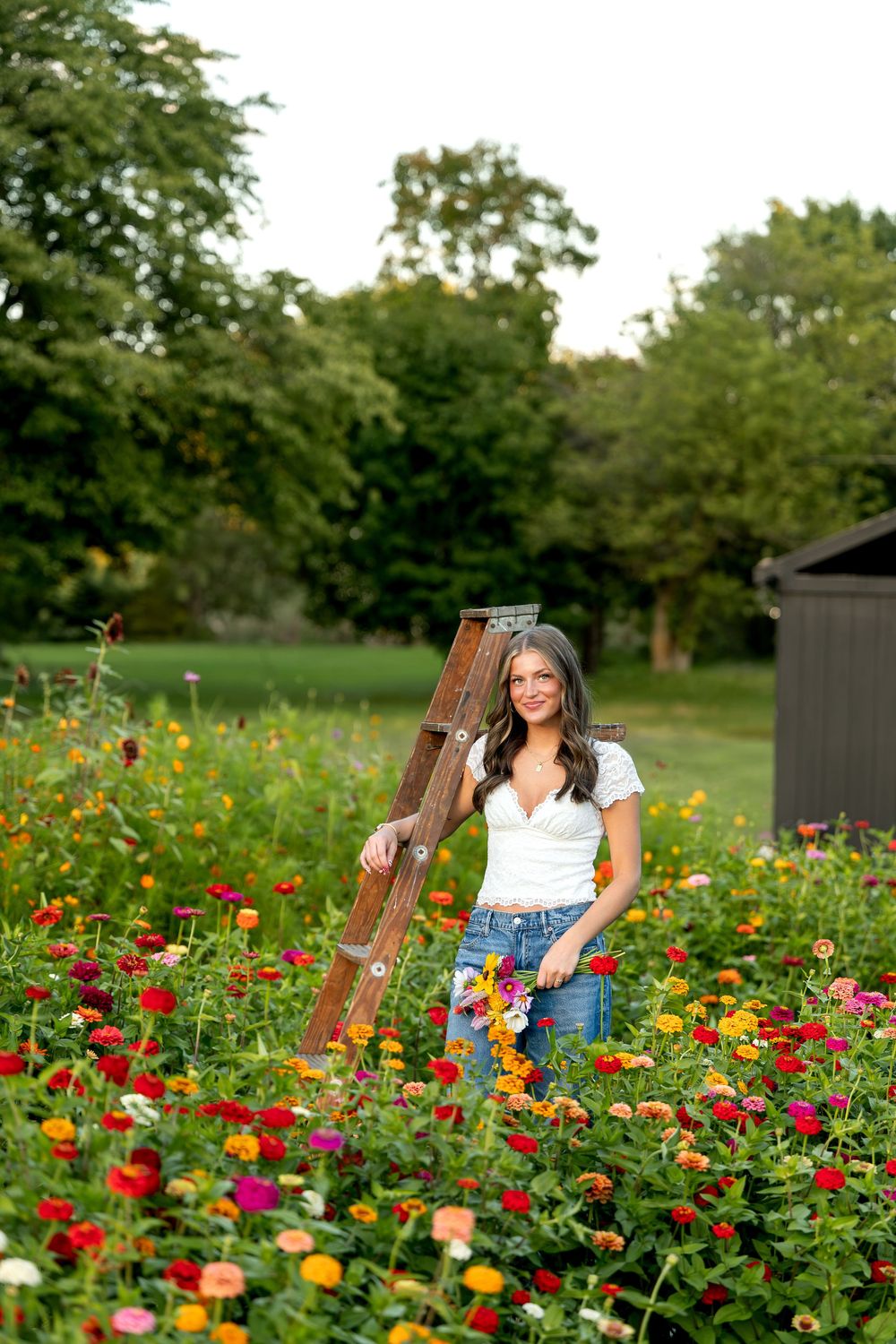 Young woman in white top and jeans smiles among colorful zinnia flowers beside a wooden ladder in a garden.