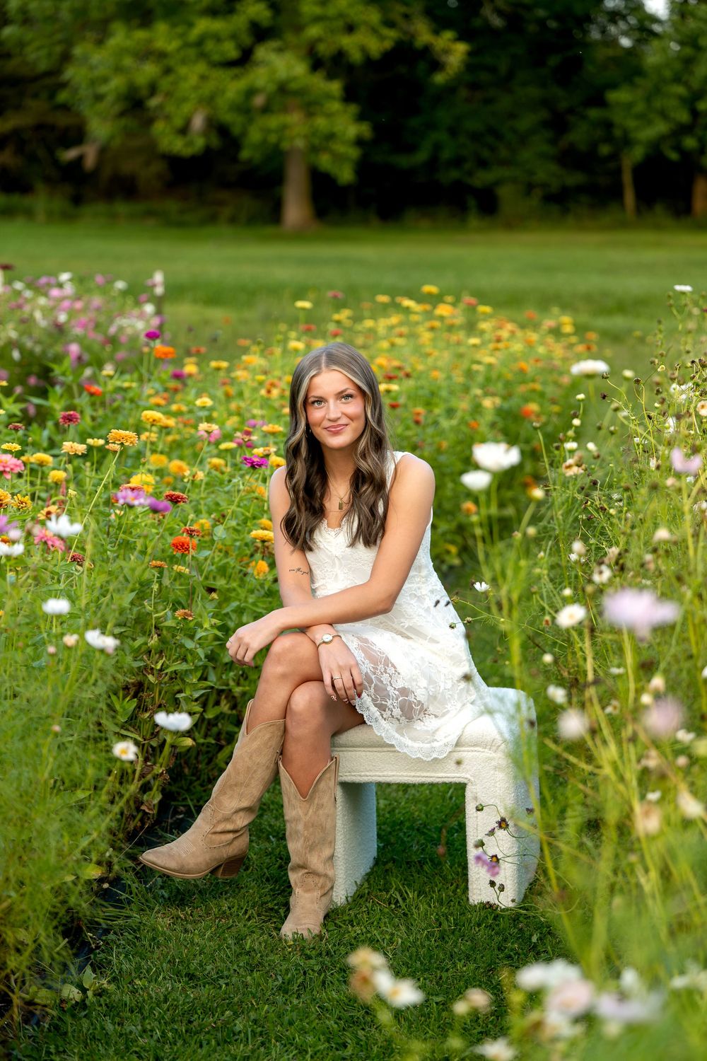 Young woman in white lace dress and cowboy boots sitting on bench surrounded by colorful wildflowers in garden.