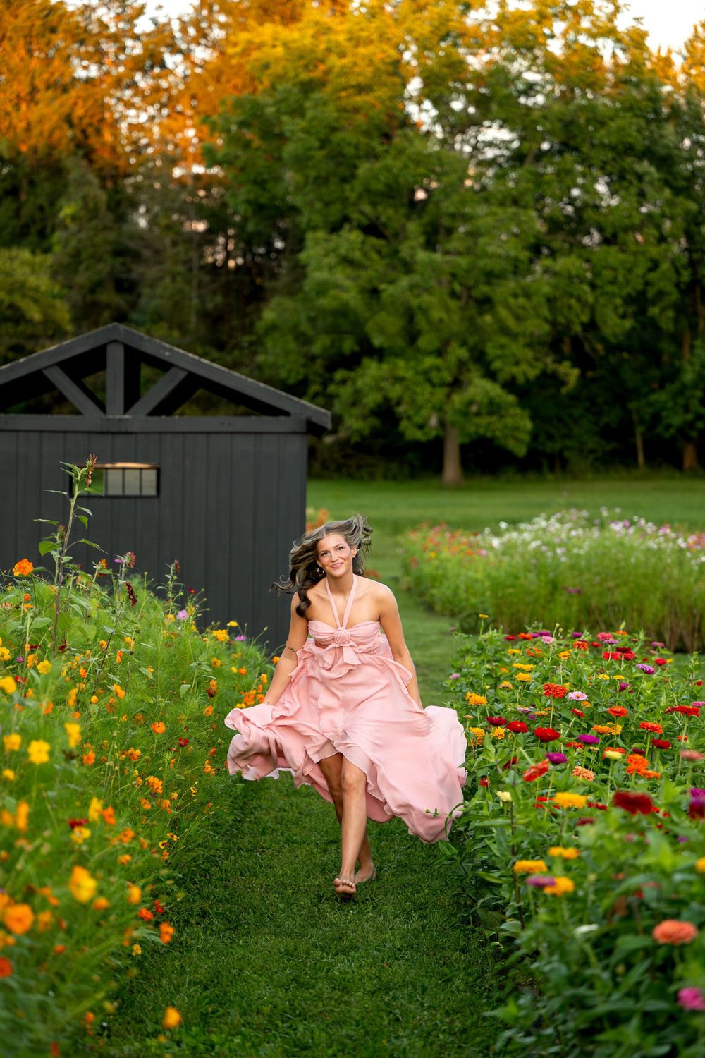 Young woman in flowing pink dress running joyfully along a garden path lined with colorful zinnias at sunset.