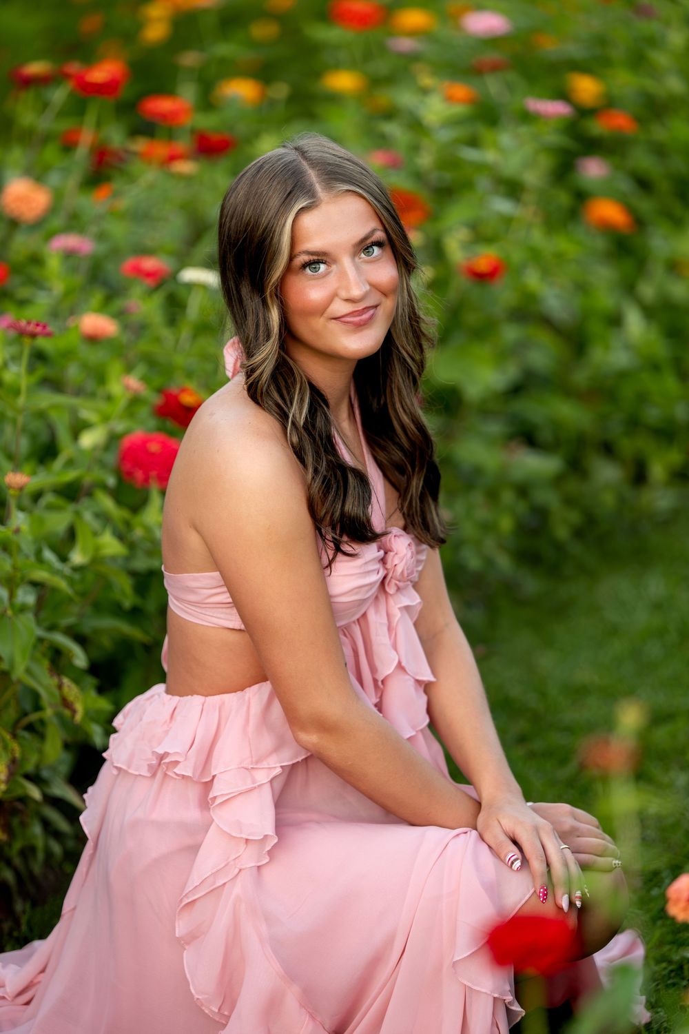 Beautiful young woman in pink ruffled dress sitting gracefully among vibrant red and orange zinnia flowers.