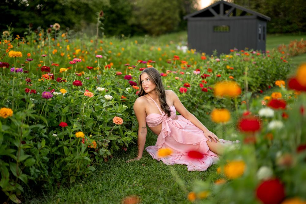 Young woman in pink dress sitting gracefully among vibrant colorful zinnias in a lush garden near a shed.