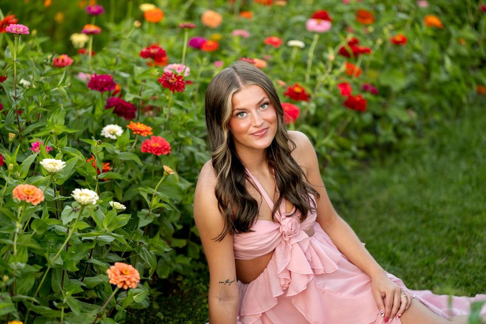 Young woman in pink dress sitting among colorful zinnia flowers in a lush garden, smiling softly.