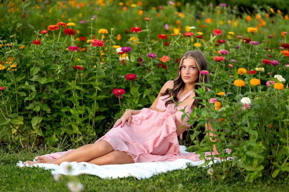 Young woman in pink ruffled dress reclining on white blanket surrounded by blooming multicolored zinnia flowers outdoors.