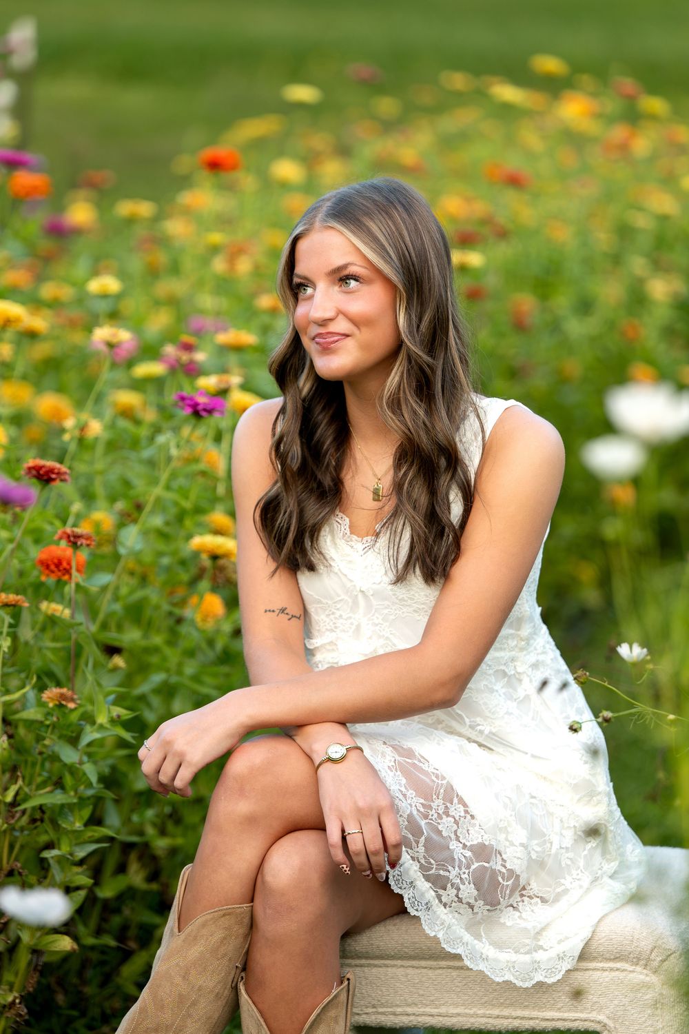 Young woman in white lace dress and cowboy boots sitting among colorful wildflowers in a sunny garden.