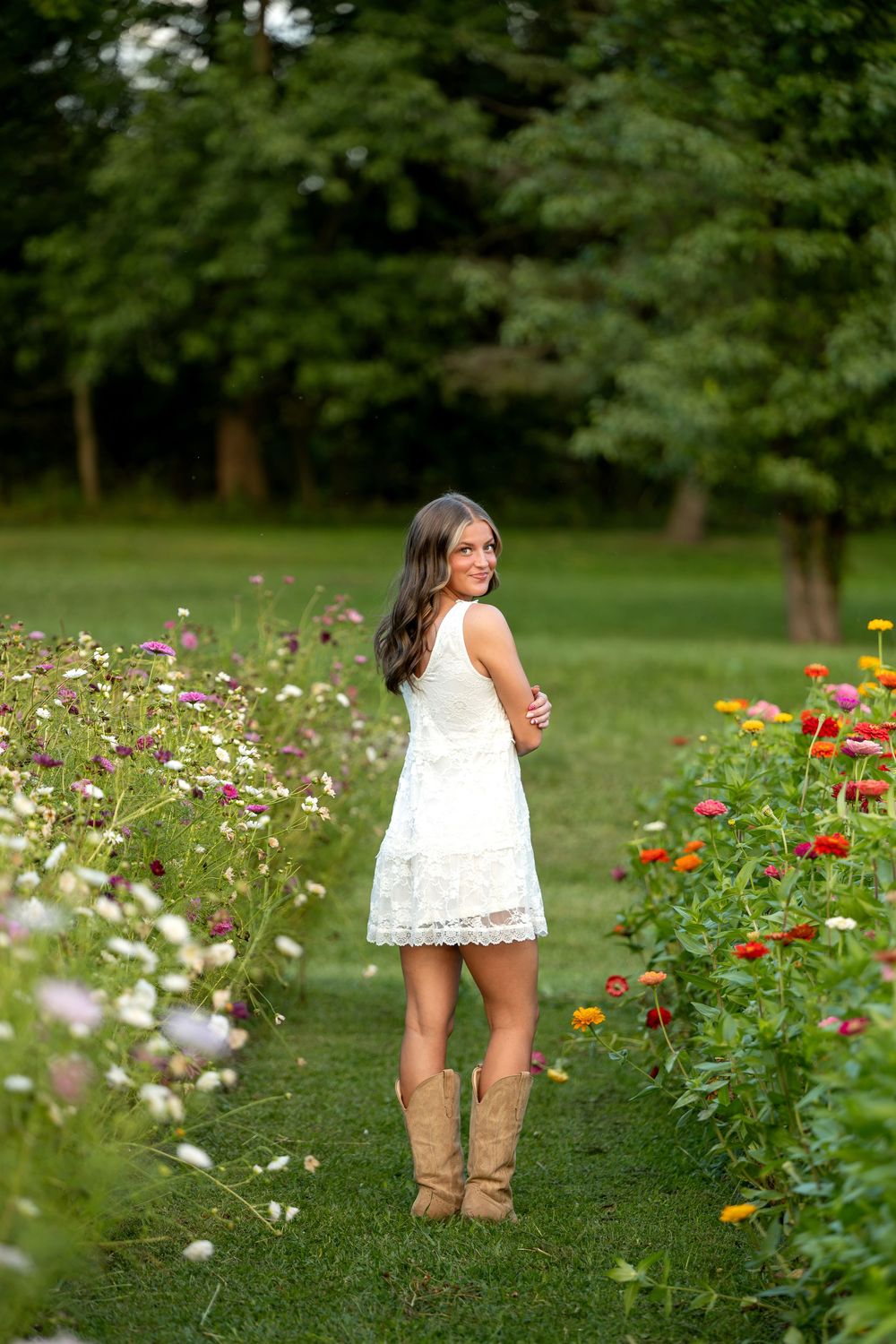 Young woman in white lace dress and cowboy boots smiling in a colorful flower garden path.