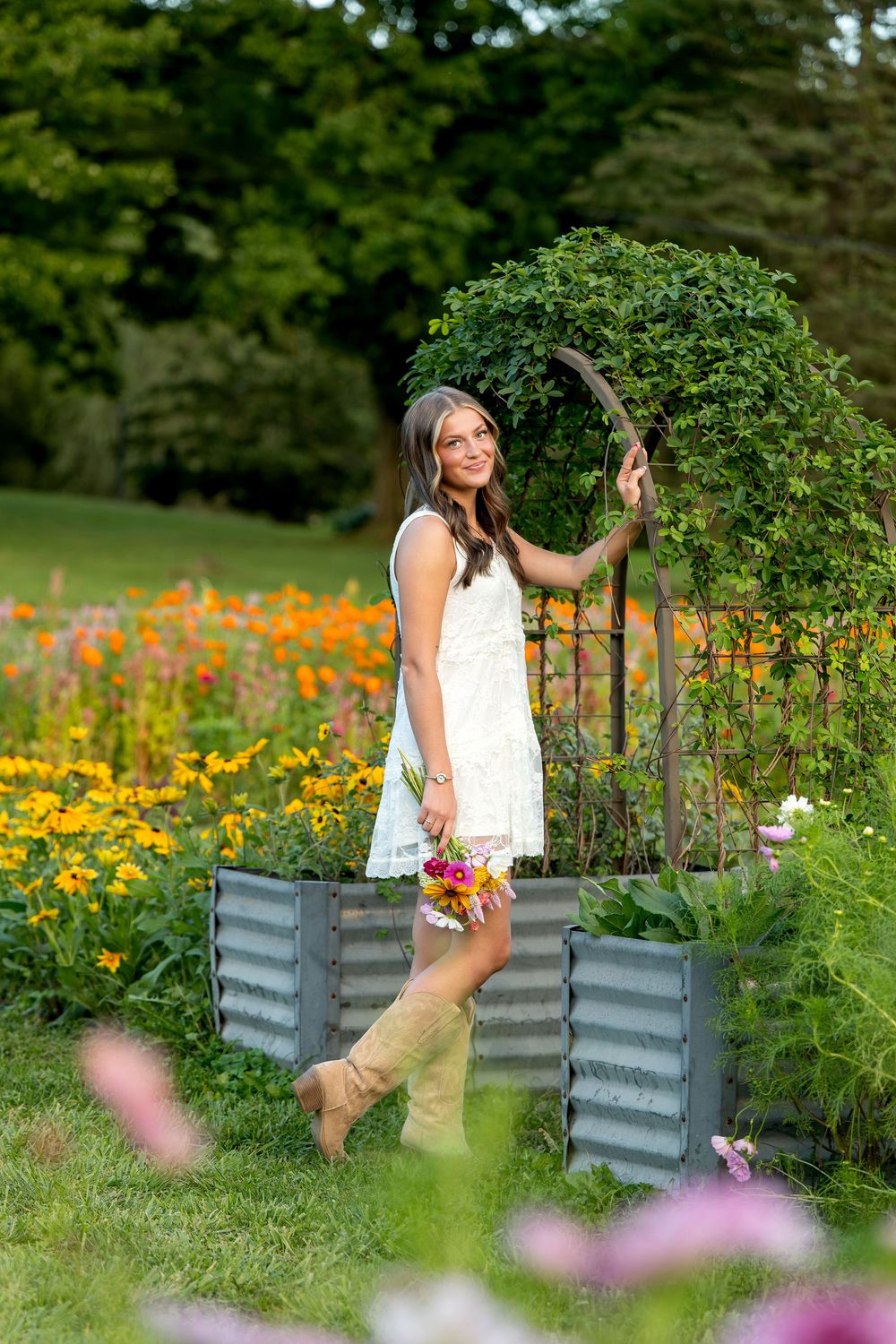 Young woman in white dress and tan boots holds flowers in a vibrant garden with raised metal beds and arch trellis.
