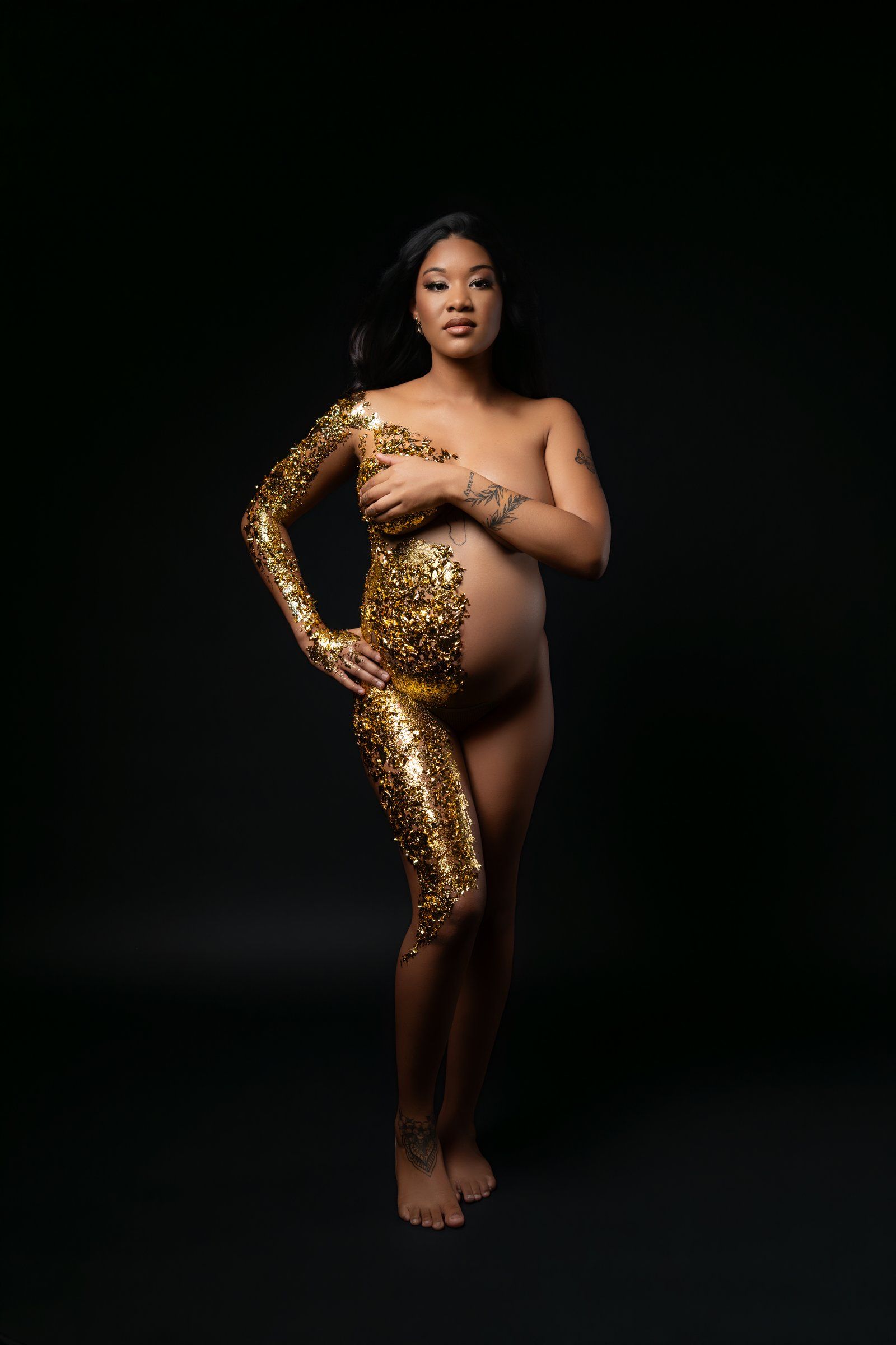 Pregnant woman posed elegantly with gold sequin body paint against a dark studio background.