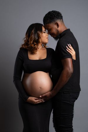A pregnant Black couple in matching black outfits share a tender moment, foreheads touching, as they cradle her baby bump.