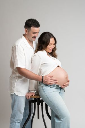 A couple poses in a studio, the man placing hands on his smiling pregnant partner's bare belly.
