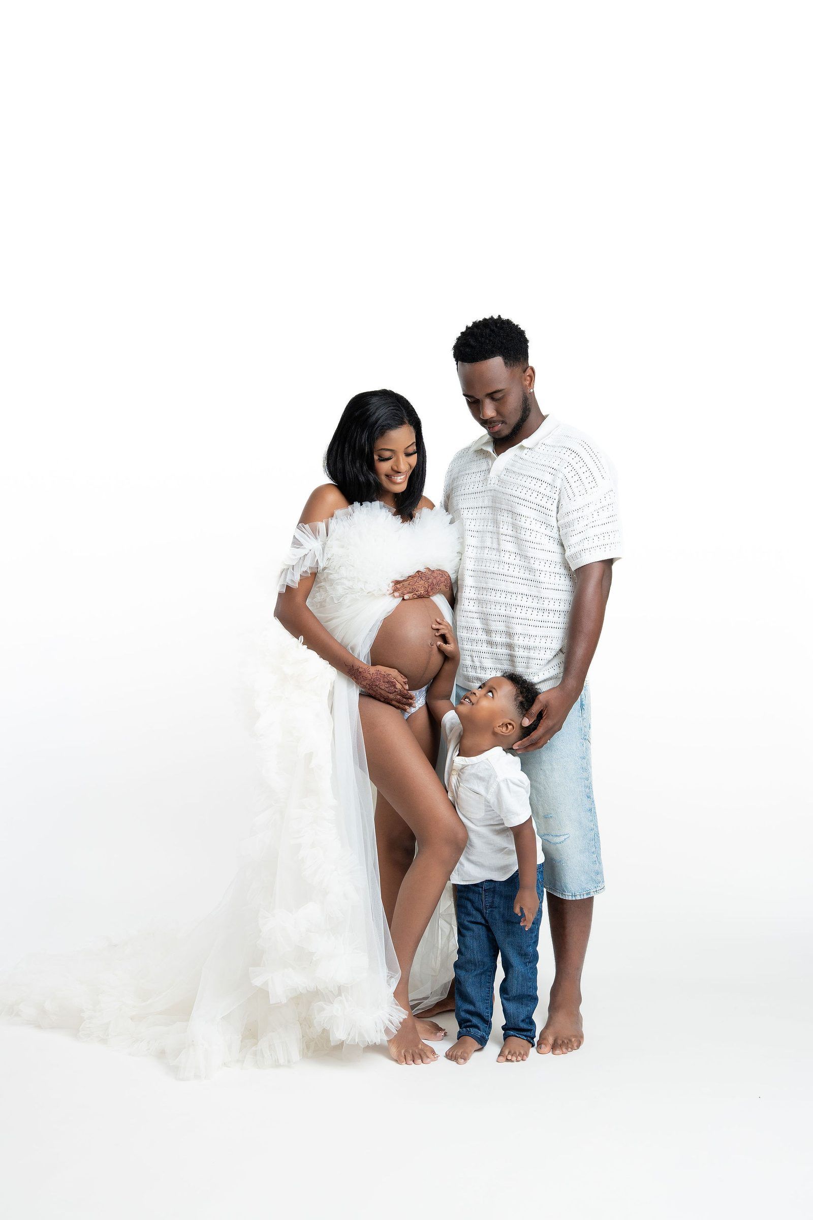 A pregnant Black woman in a white ruffle gown poses with her partner and young child in a studio maternity photo.
