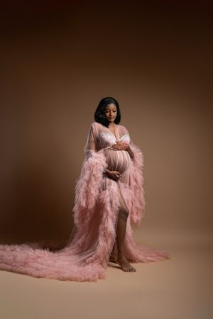 Pregnant woman in flowing pink tulle robe cradling baby bump against warm brown studio background.
