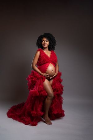 Pregnant woman in red ruffled maternity gown cradling baby bump, studio portrait photography.