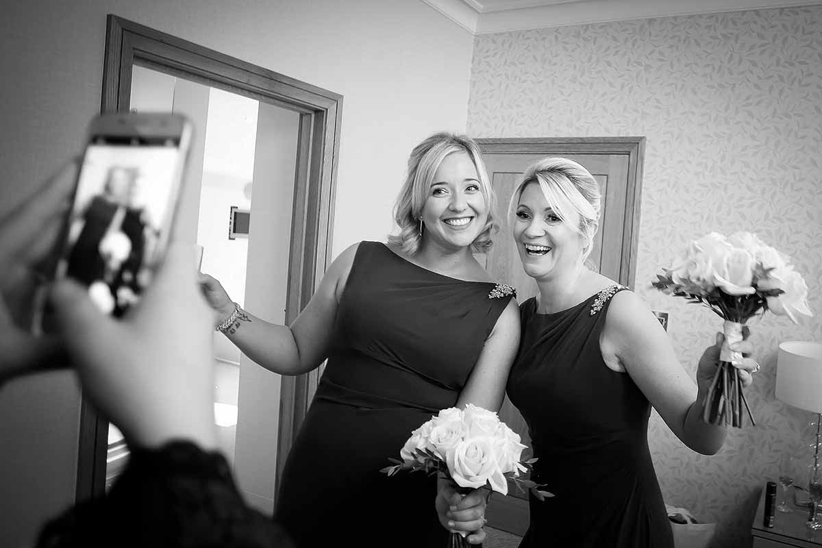 Two bridesmaids in black dresses holding white bouquets pose for a photo in black and white wedding preparation shot.