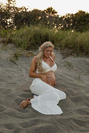 Pregnant woman in white outfit sitting on sandy beach dunes at sunset, cradling her baby bump.