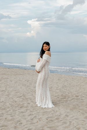 Pregnant woman in white lace off-shoulder gown poses on sandy beach, smiling over shoulder with ocean backdrop.