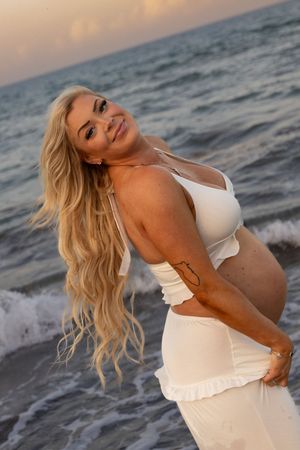 Smiling pregnant woman with long blonde hair poses in white crop top and skirt on beach at sunset.