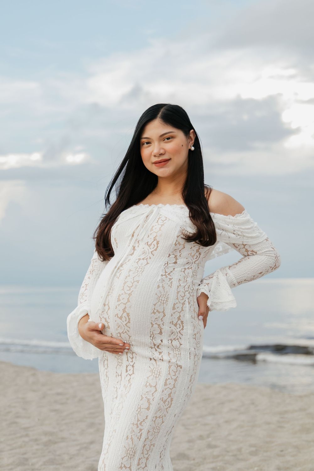Pregnant woman in white lace off-shoulder gown posing on a sandy beach with cloudy sky backdrop.