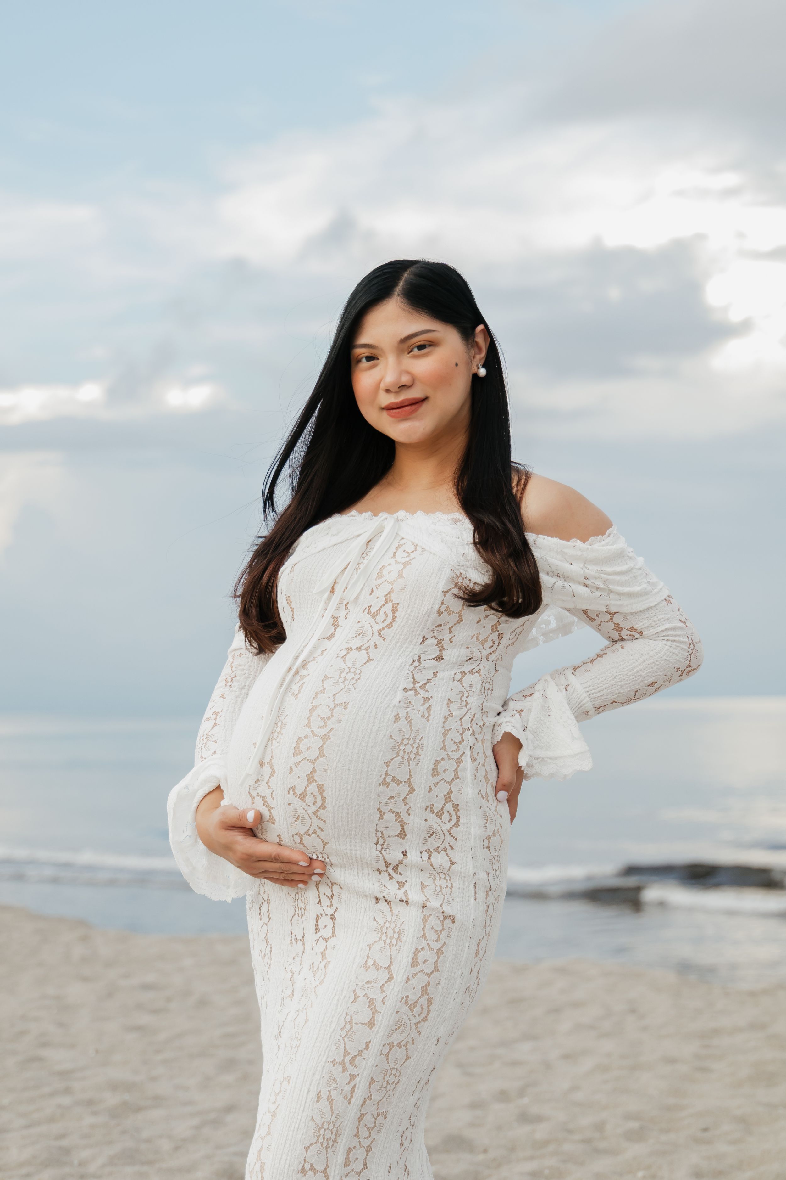Pregnant woman in white lace off-shoulder gown posing on a sandy beach with cloudy sky backdrop.