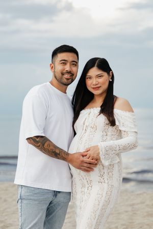 A smiling couple poses on a beach, the man in a white tee with tattoos embraces a pregnant woman in a white lace dress.