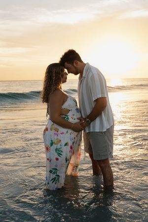 Couple standing in ocean waves at sunset, holding hands and touching foreheads romantically on a tropical beach.