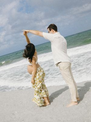 Couple dancing on a sandy beach with ocean waves, man spinning woman in floral skirt on a sunny day.