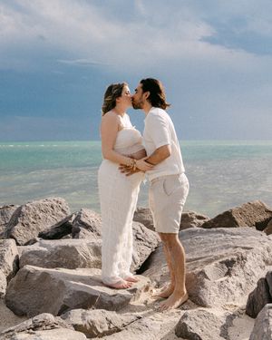 Couple kissing on rocky shoreline, woman in white maternity gown, man in white outfit, ocean backdrop.