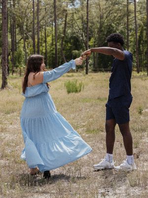 A couple dances together in a sunlit forest clearing, the woman wearing a flowing light blue off-shoulder dress.