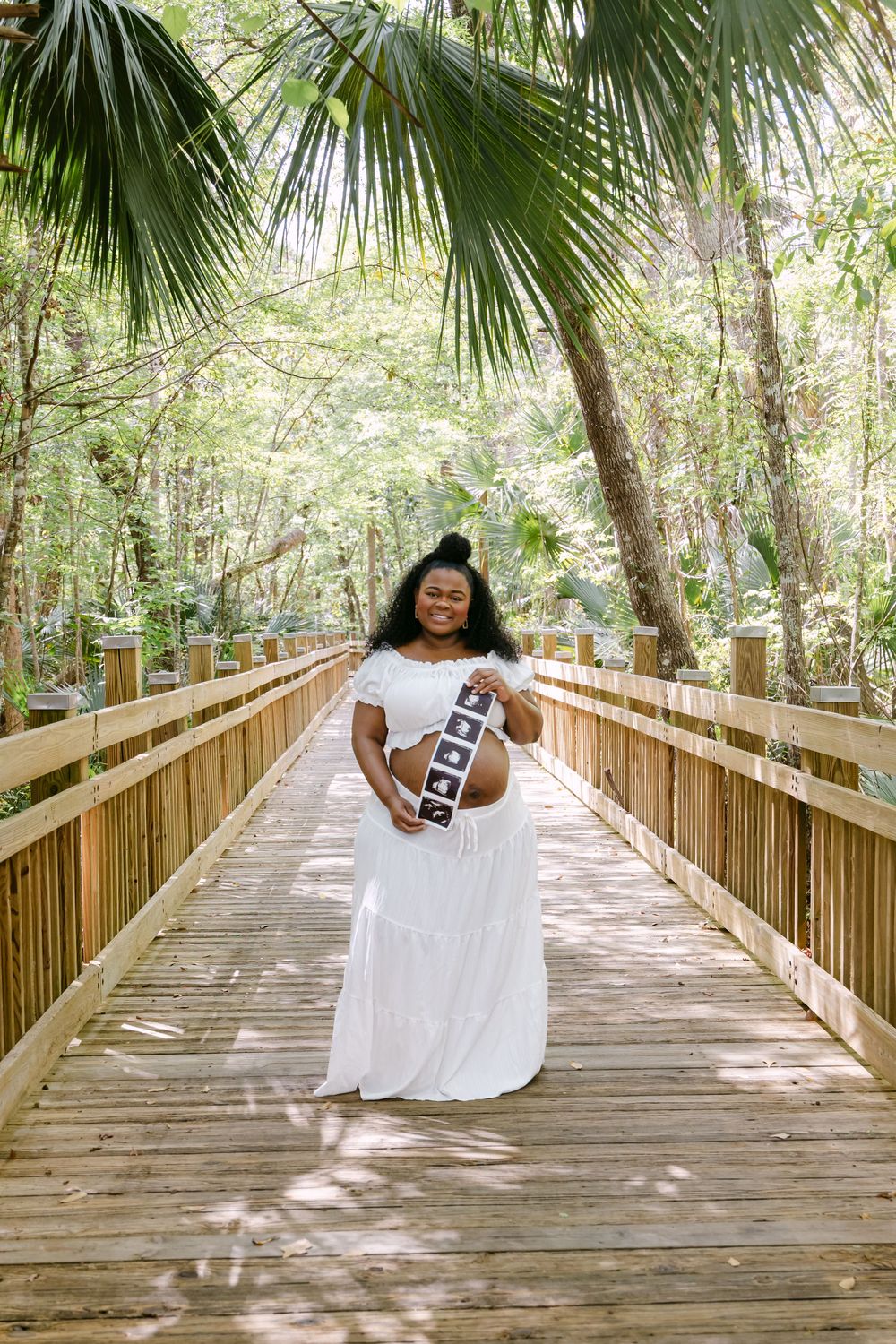 Pregnant woman in white off-shoulder dress holds ultrasound photos on a sunny wooden boardwalk surrounded by lush greenery.