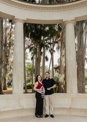 Couple holding ultrasound photos at elegant columned rotunda surrounded by palm trees, announcing pregnancy.