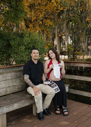 Expecting couple sits on park bench holding baby onesie and ultrasound photos, surrounded by autumn trees near a pond.