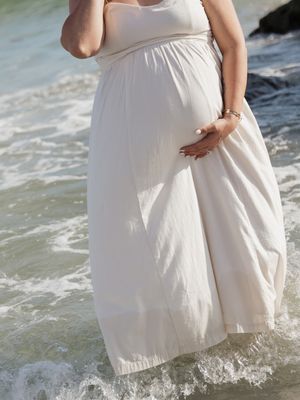 Pregnant woman in white maxi dress standing in ocean waves at beach, cradling baby bump.