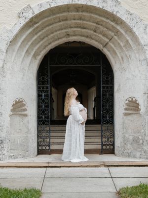 Pregnant woman in white lace gown standing under ornate stone arch doorway with black iron gates.
