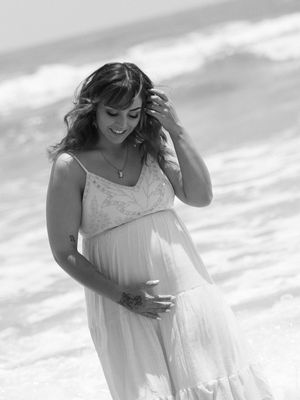 Pregnant woman in white dress at beach, smiling down at baby bump, black and white maternity photo.