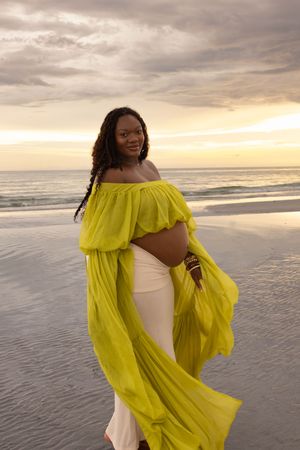 Pregnant woman in yellow off-shoulder dress poses on beach at sunset, showcasing baby bump.