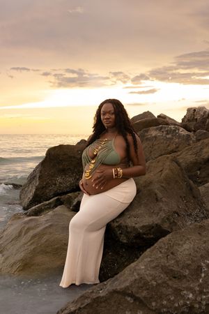 Pregnant woman in green top and white skirt poses on rocks at sunset beach, cradling her baby bump.