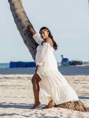 Woman in white off-shoulder maxi dress posing by a palm tree on a sandy beach.