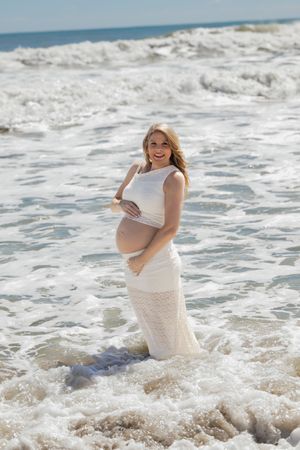 Smiling pregnant woman in white lace dress standing in ocean waves on a sunny beach, cradling her baby bump.