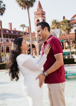 Pregnant couple smiling at each other near a fountain with a historic red building and palm trees in the background.