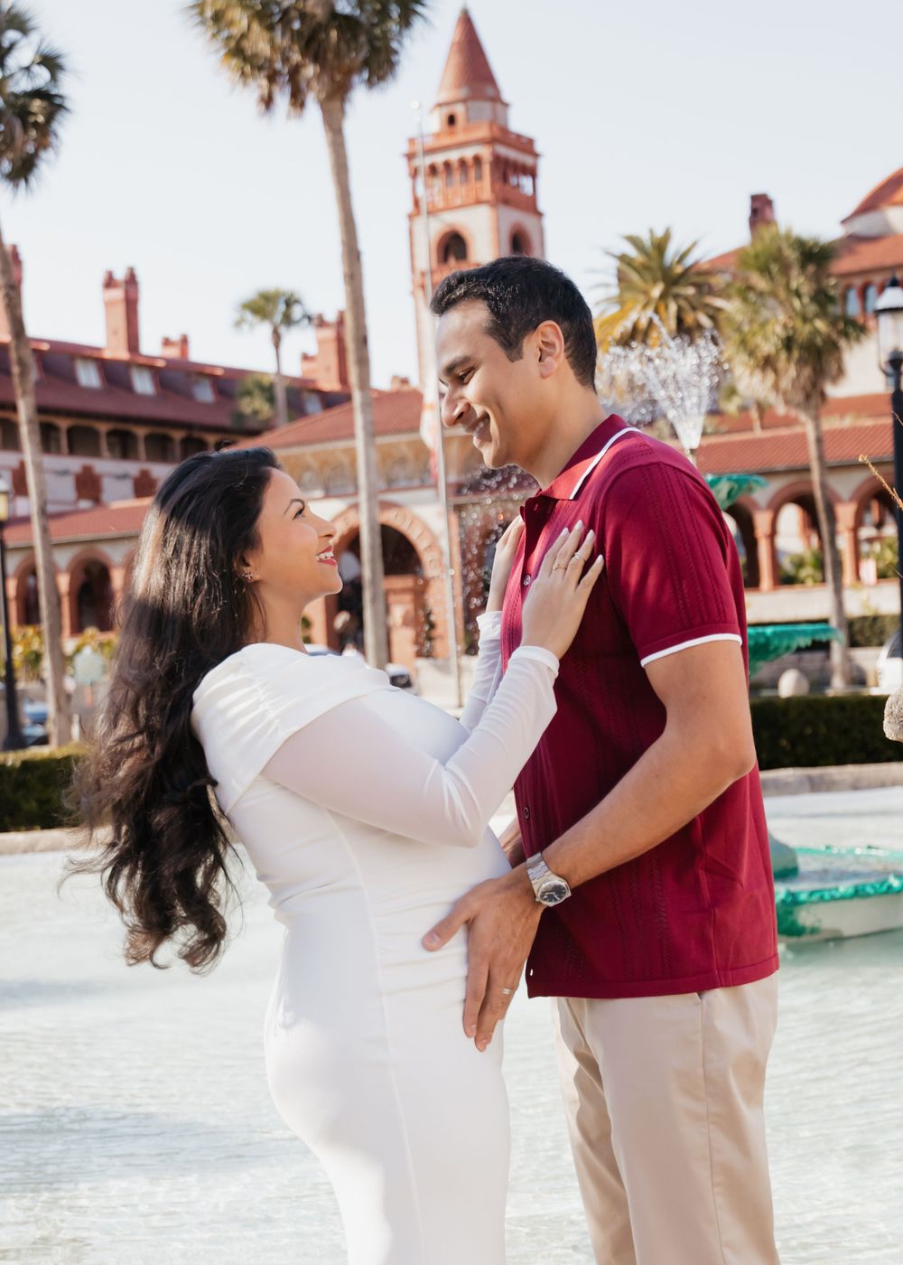 Pregnant couple smiling at each other near a fountain with a historic red building and palm trees in the background.