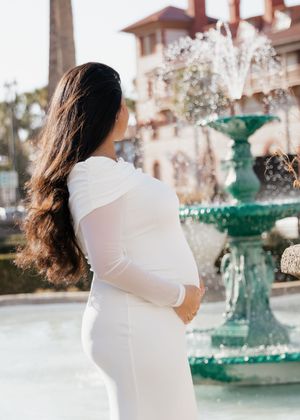 Pregnant woman in white dress holding baby bump near green fountain outdoors.